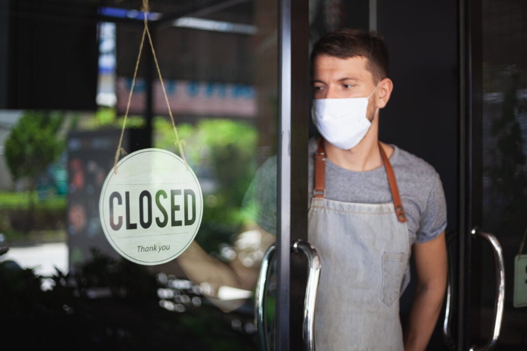Chef wearing face mask hanging closed sign on restaurant door.